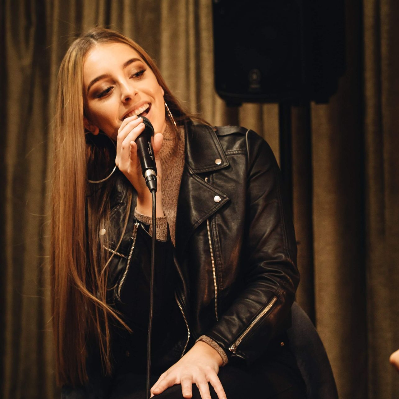 A person with long hair singing into a microphone, wearing a black leather jacket in front of brown curtains.