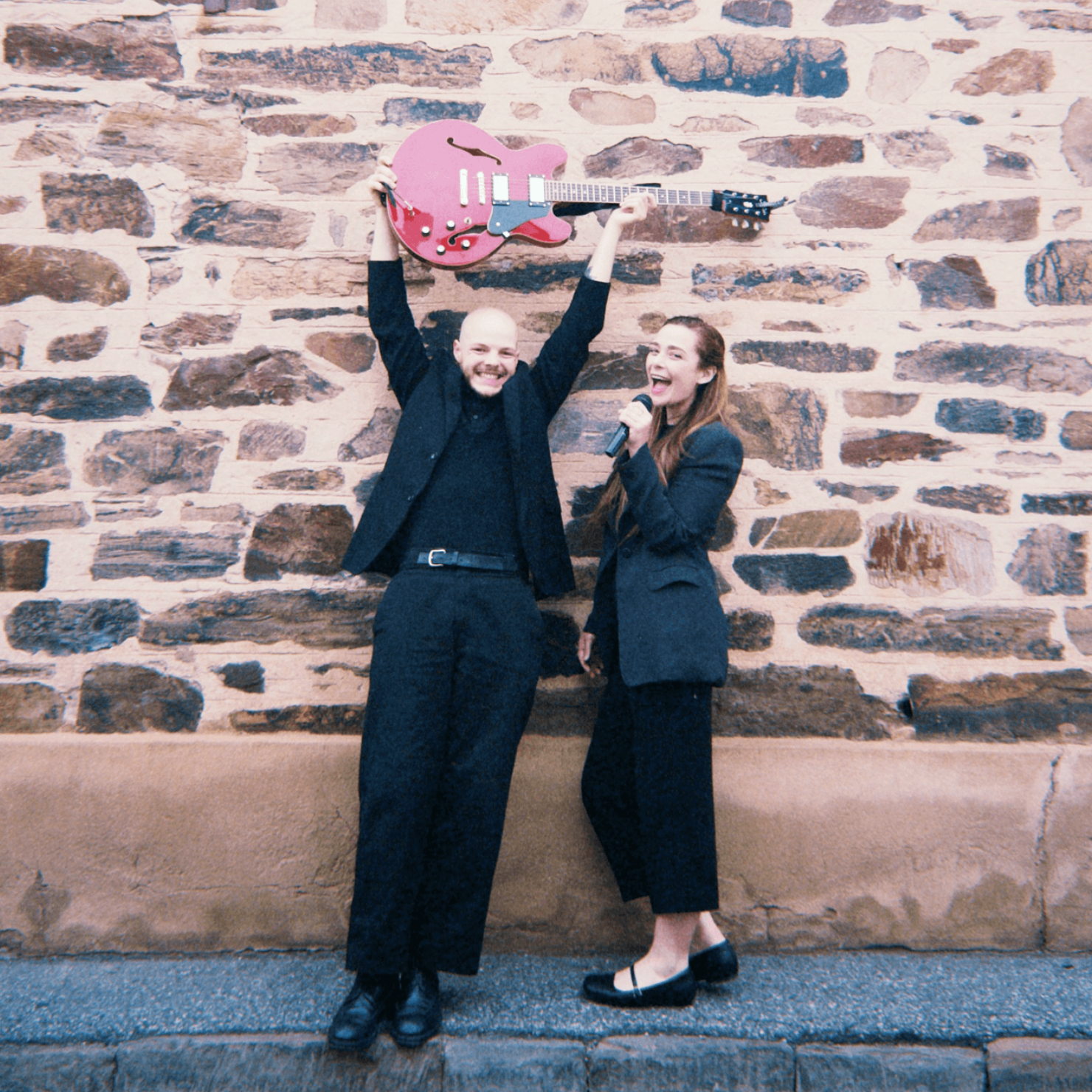 Two people outdoors by a stone wall, one lifting a pink electric guitar overhead while the other laughs.