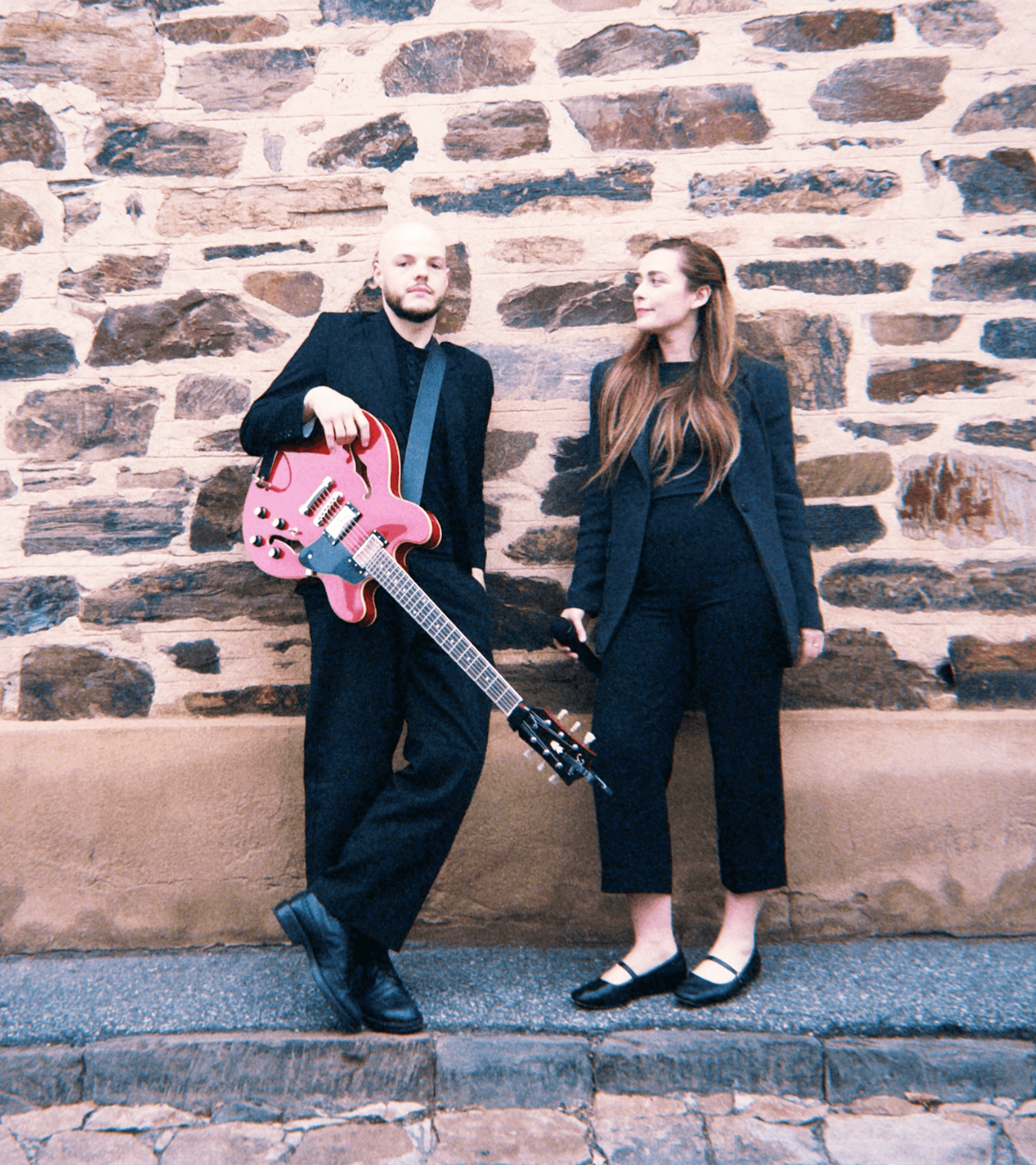 Two people in black outfits stand against a stone wall, one holding a pink guitar.