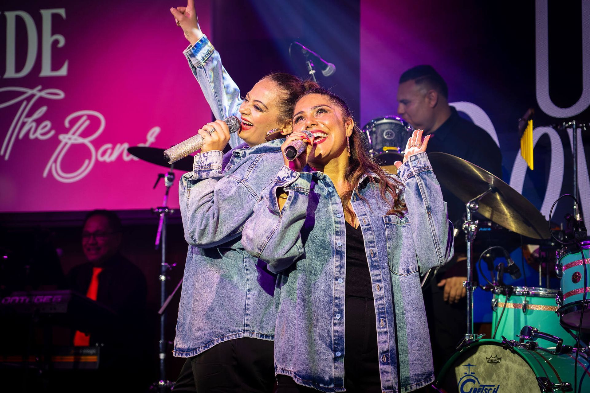 Two singers in silver jackets performing on stage with colorful lights and a drummer behind them