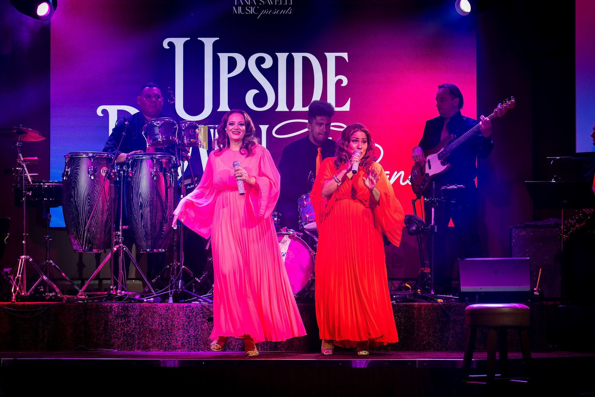 Two singers in pink and red gowns perform onstage under purple lights with band and “UPSIDE” backdrop
