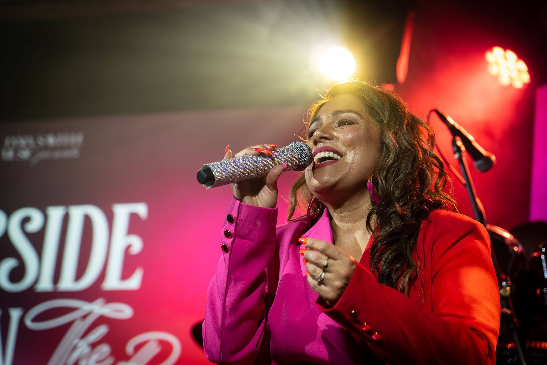 Singer in a red jacket performing on stage with a microphone under red lights