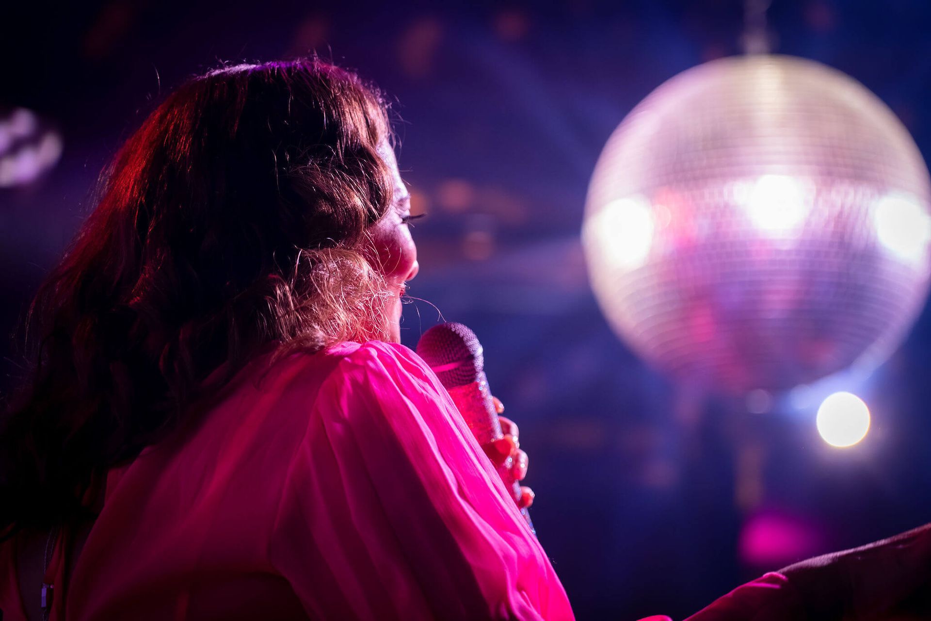 Person in a pink outfit singing into a microphone under purple stage lights with a disco ball overhead