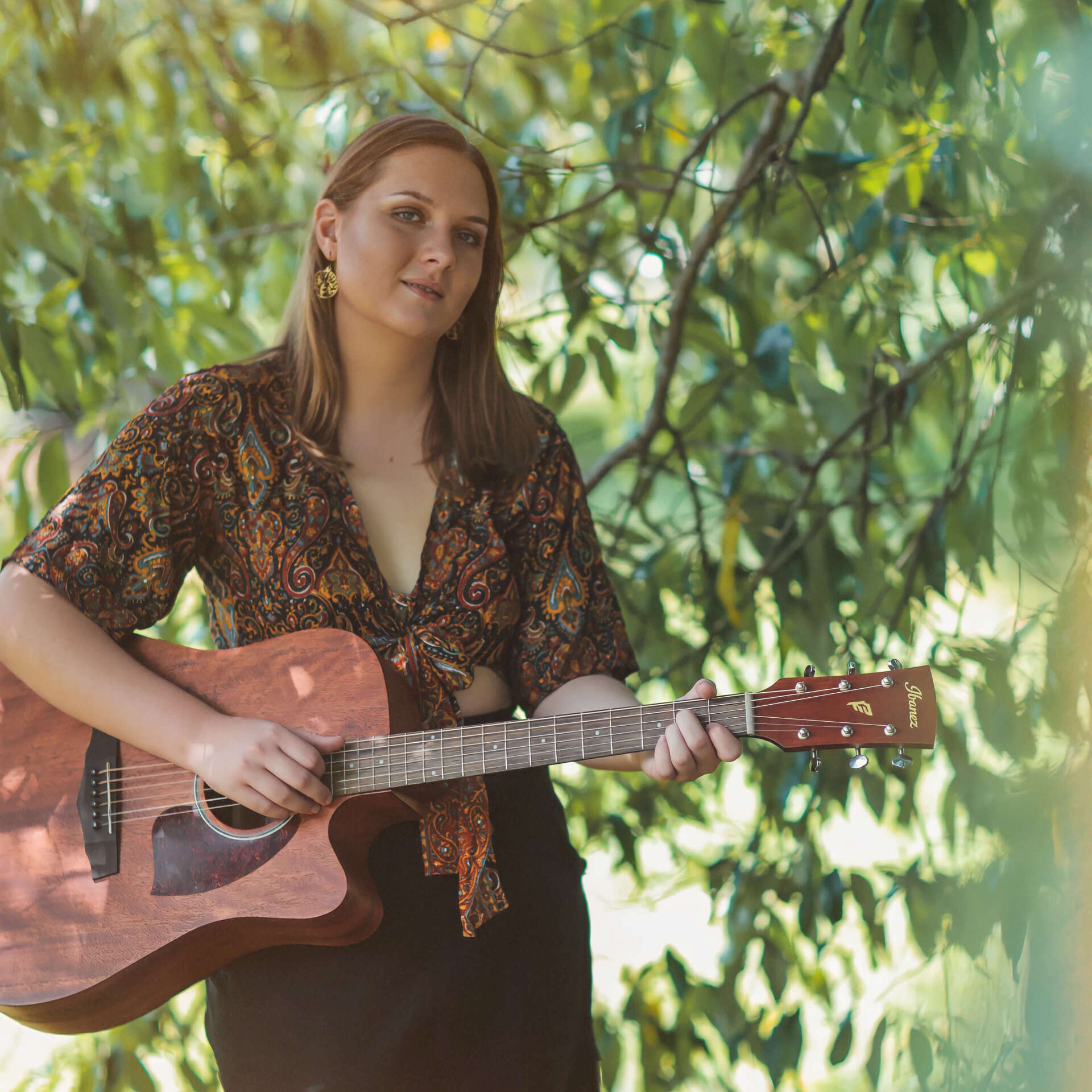 A person with long hair wears a patterned top while playing an acoustic guitar among lush green tree branches.