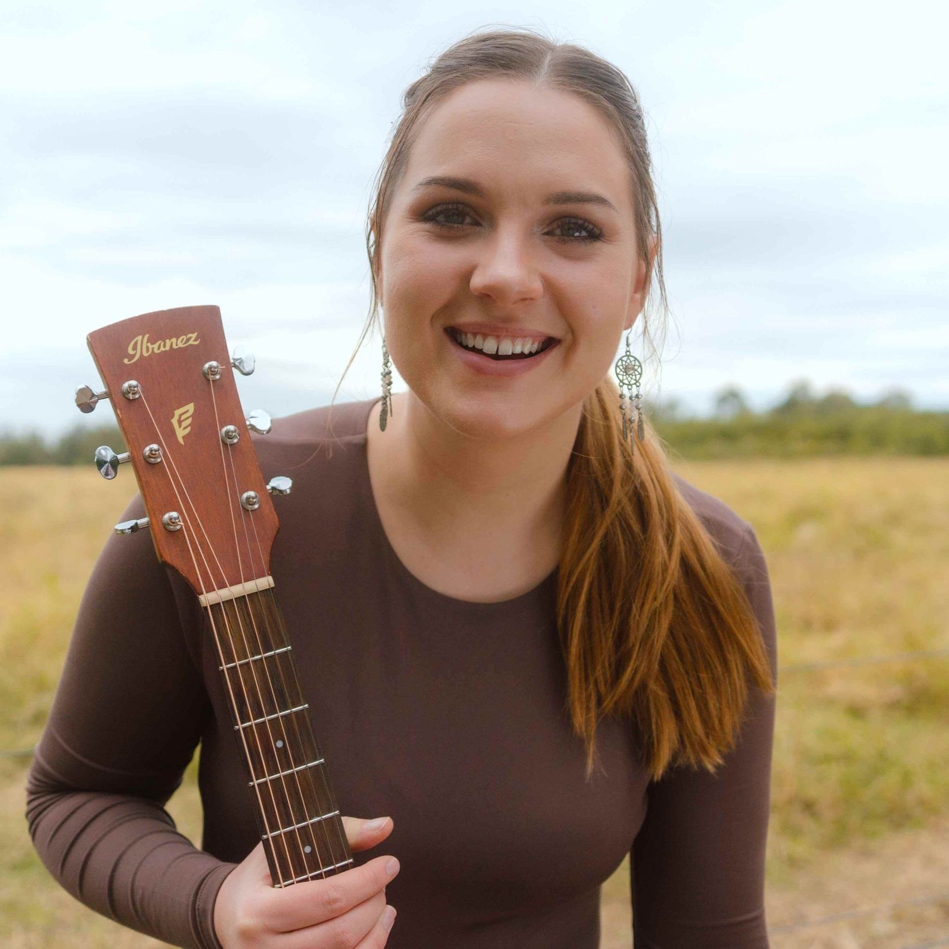 A person smiling while holding an Ibanez acoustic guitar in a field.
