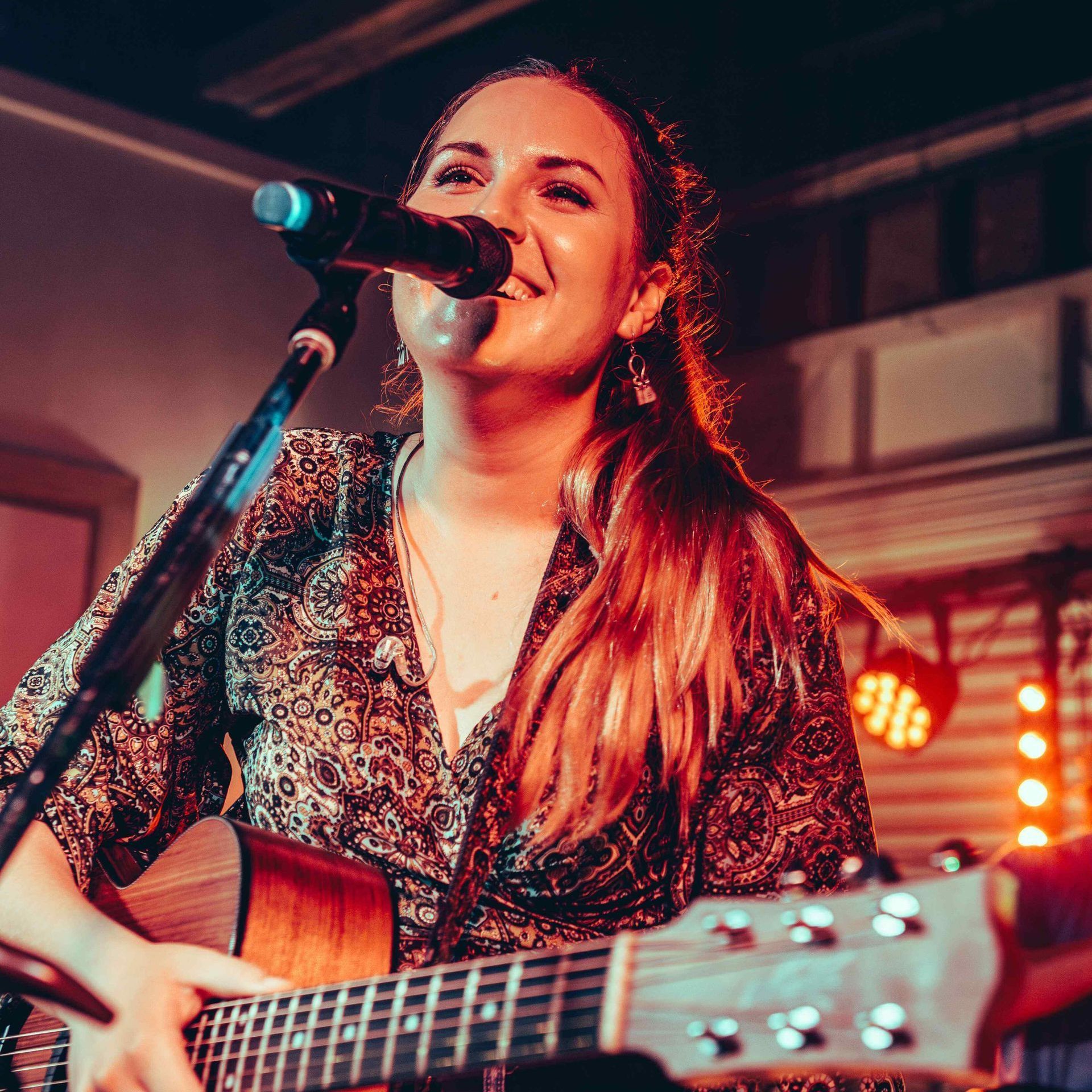 A smiling performer plays an acoustic guitar and sings into a microphone on a stage with warm, glowing stage lights.