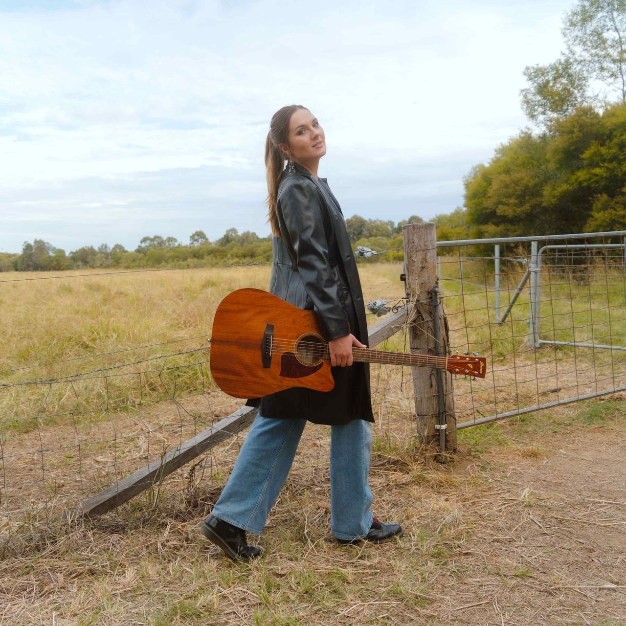 A person holding an acoustic guitar walks past a wooden fence in a rural field under a bright, overcast sky.