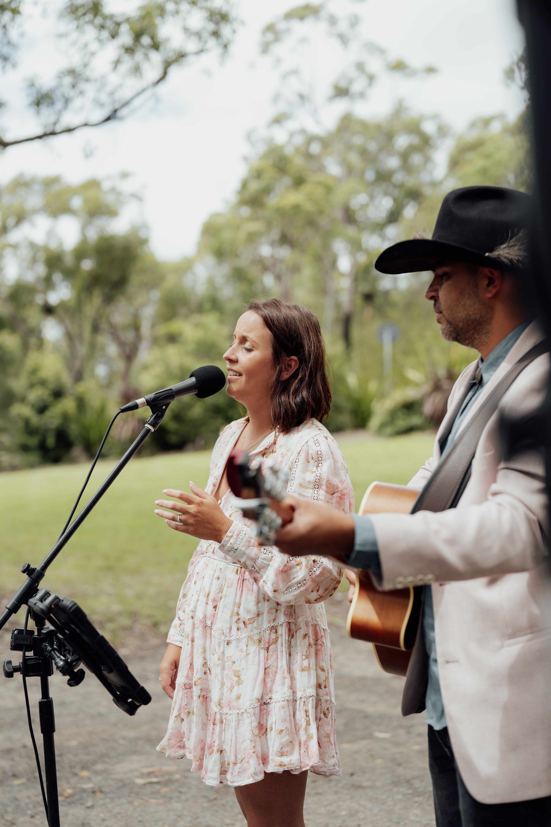 Outdoor live music performance with a singer at a microphone and guitarist beside her in a park