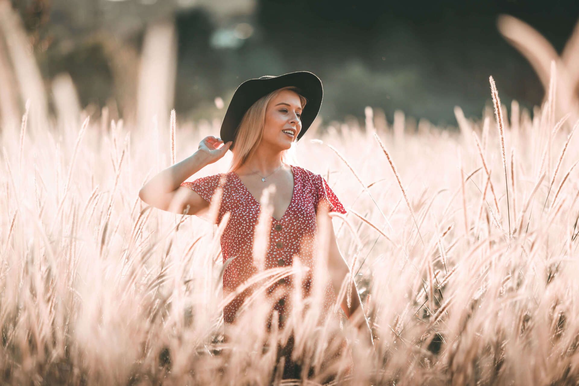 A person wearing a wide-brimmed hat and patterned top smiling while standing in a sunlit field of tall grass.