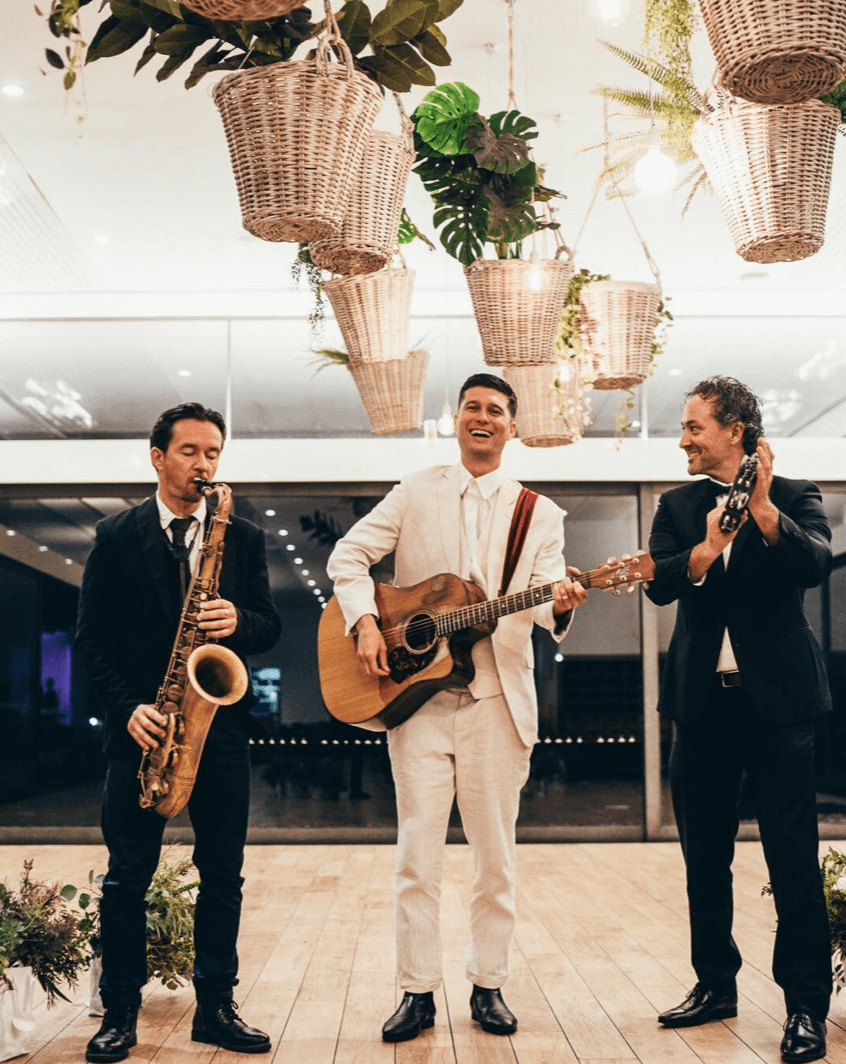 Three musicians perform indoors, with a saxophonist and a guitarist playing and a third person clapping, under decor.