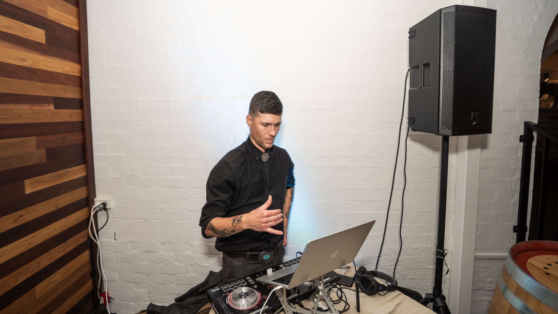 A person in a black shirt DJs at a laptop and mixing board, standing in front of a white wall next to a tall speaker.