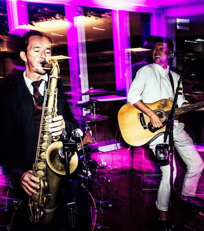 A musician plays a saxophone while another plays an acoustic guitar on a stage lit with bright purple lights.