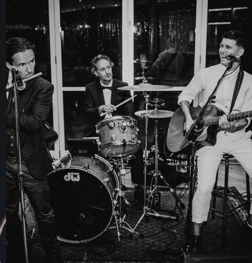 A black-and-white photo of a three-piece band performing indoors, featuring a flutist, a drummer, and a guitarist.