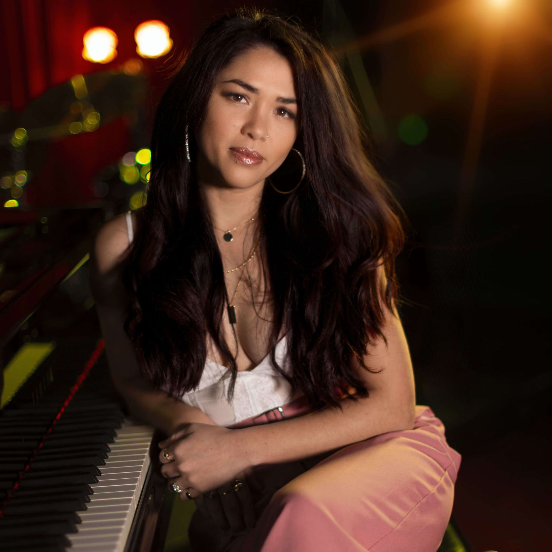 A woman with long dark hair sits by a piano, wearing a white top and pink pants, looking towards the camera with a gentle expression.