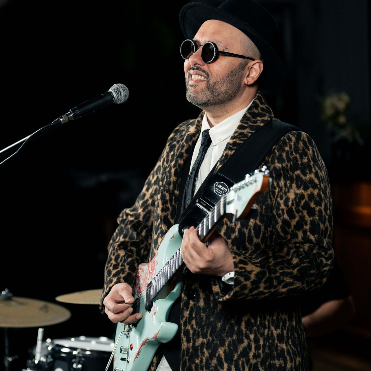 Man playing guitar, wearing leopard print coat, sunglasses, and hat, smiling at a microphone.