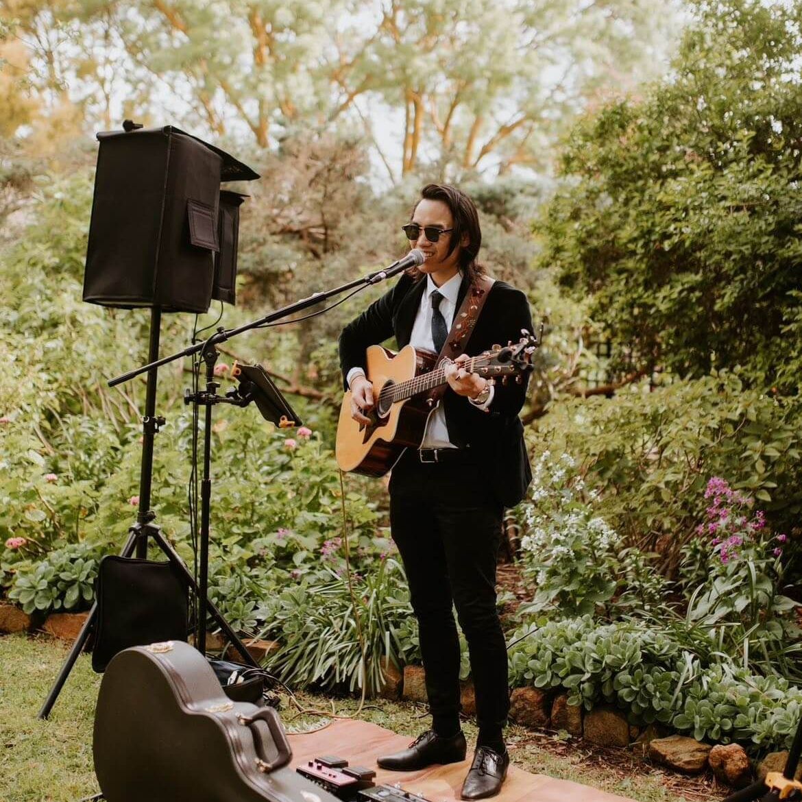 Man in black suit playing acoustic guitar outdoors, microphone, speaker, and guitar case visible.