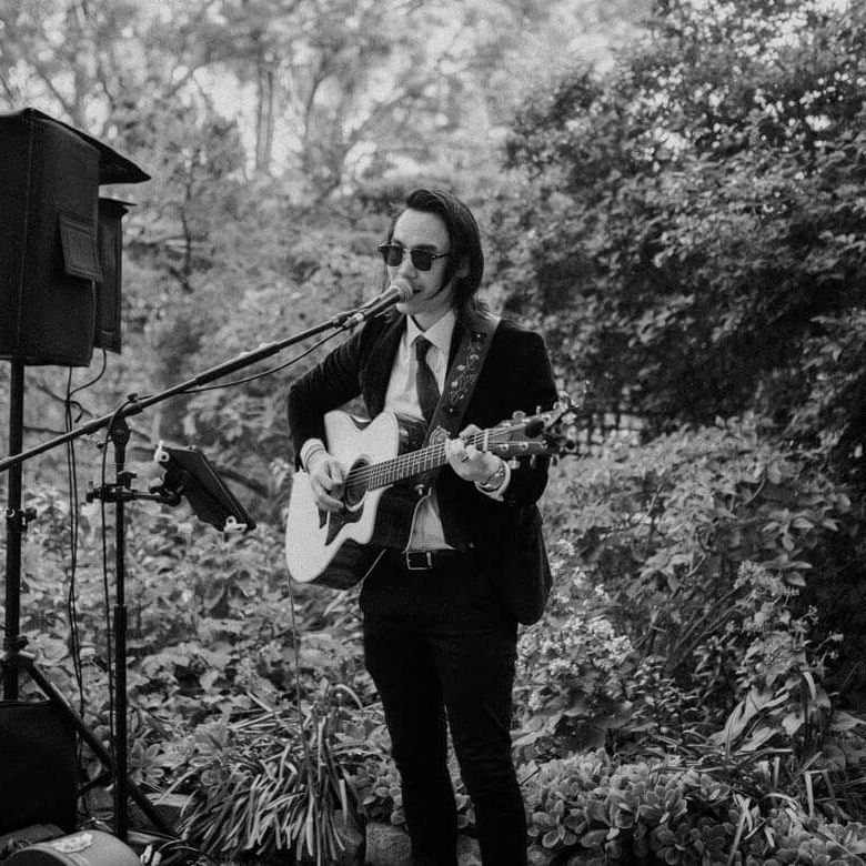 Musician singing and playing guitar outdoors at a microphone, with greenery behind him.
