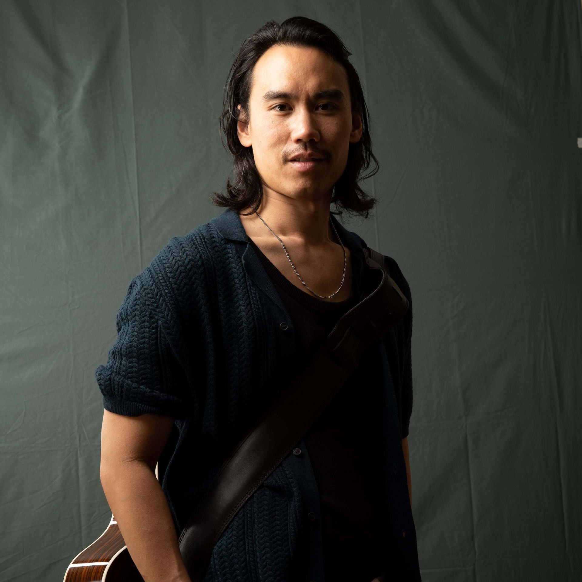 Man with long dark hair, wearing a dark blue shirt and holding a guitar, against a gray background.