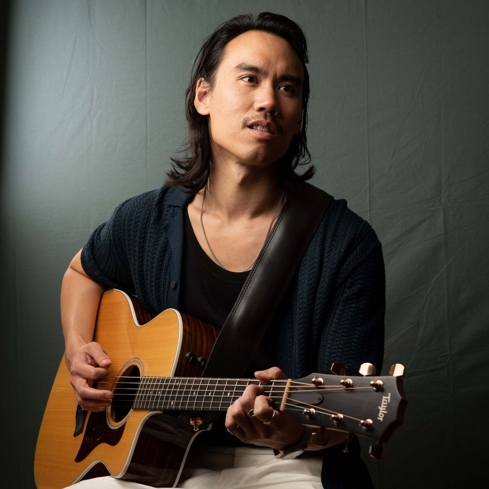 Man with long hair, playing an acoustic guitar, wearing a dark blue shirt and looking upwards.