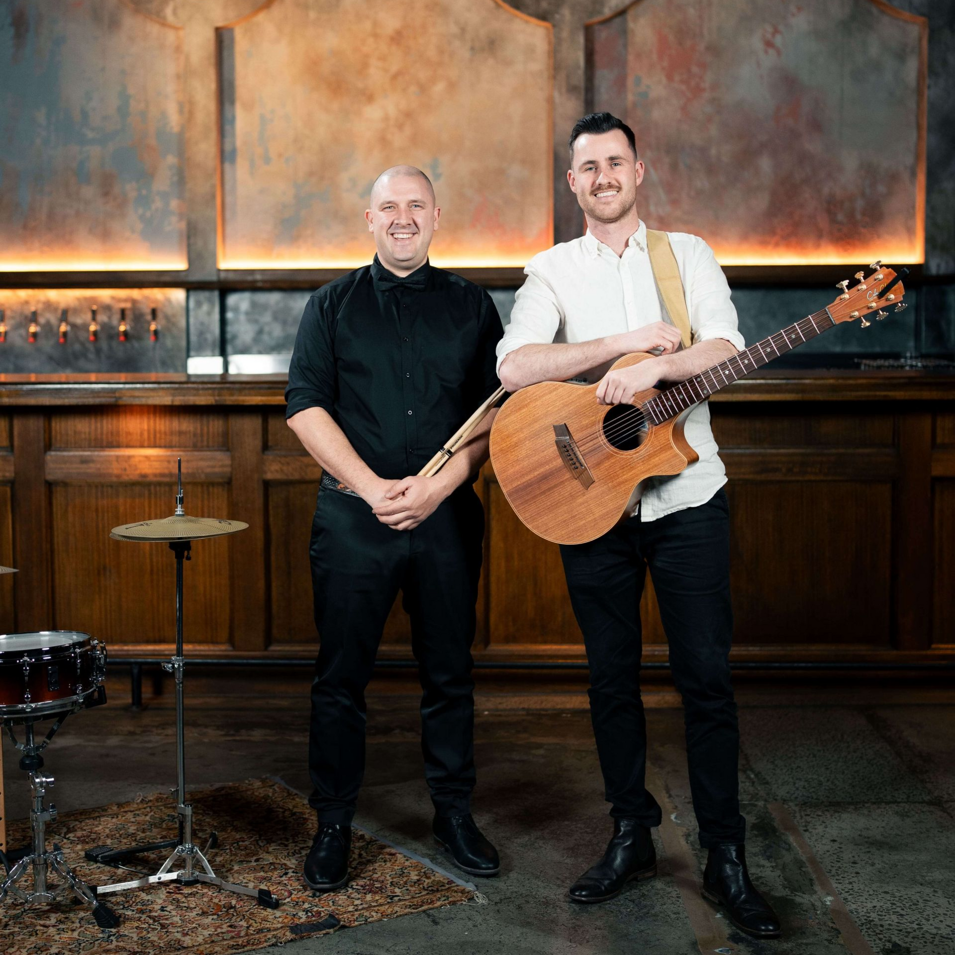 Two men in a bar. One plays guitar, the other holds drum sticks, both smiling. Warm, rustic setting.
