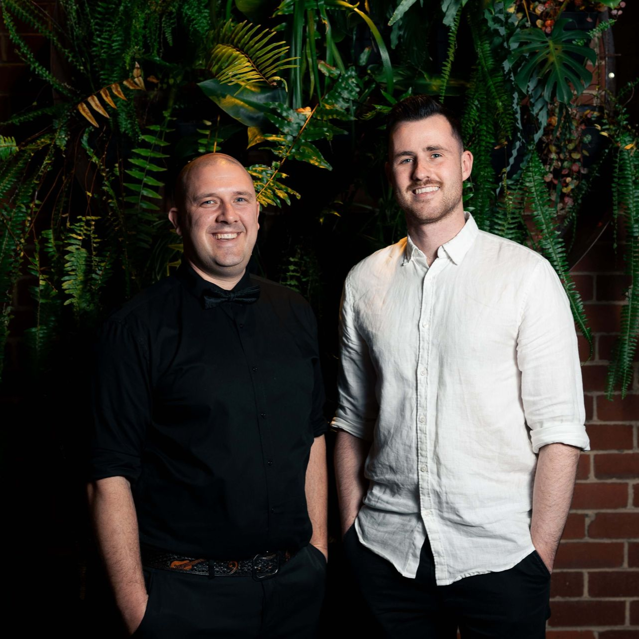 Two men pose, one in black shirt and bow tie, the other in white shirt. Behind them is greenery and a brick wall.