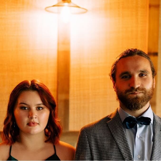 Woman and man in formal attire under a soft light. The man has a beard and bow tie.