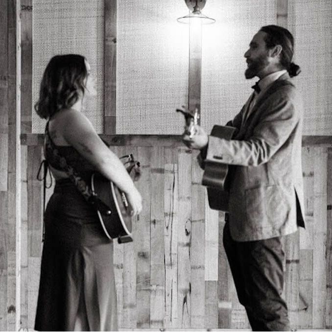 Two musicians with guitars facing each other in front of a wood-paneled wall, performing under a light.