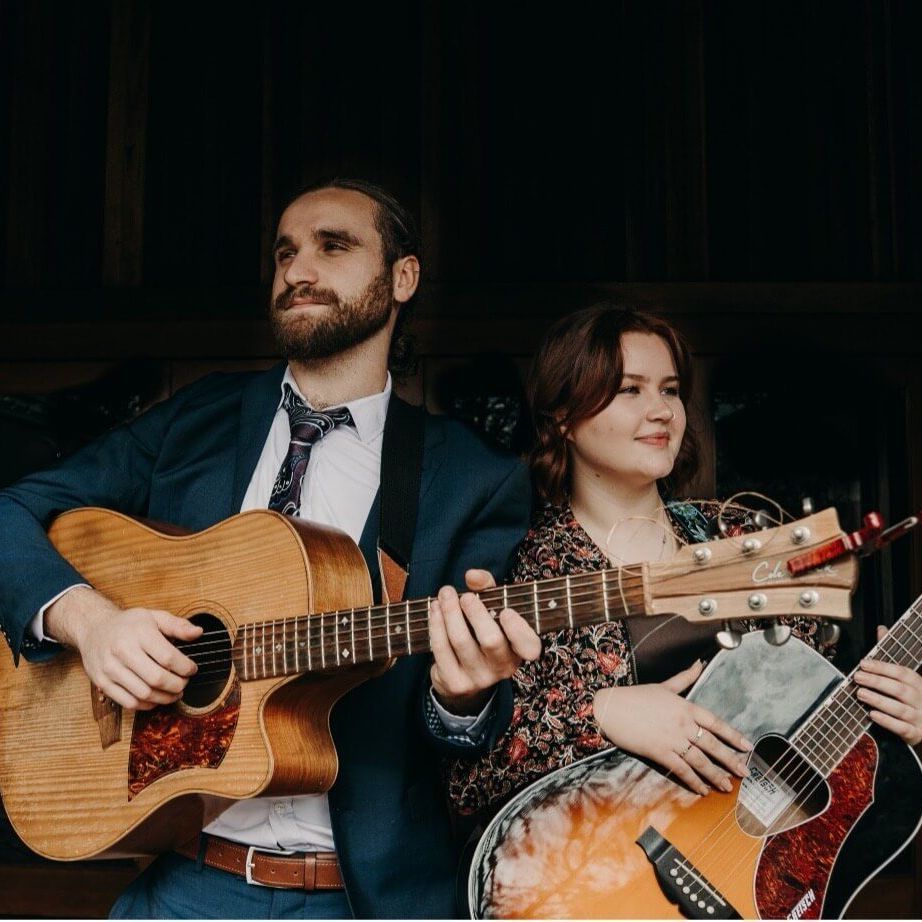 Two musicians playing acoustic guitars. Man in blue suit, woman in floral shirt, both smiling.