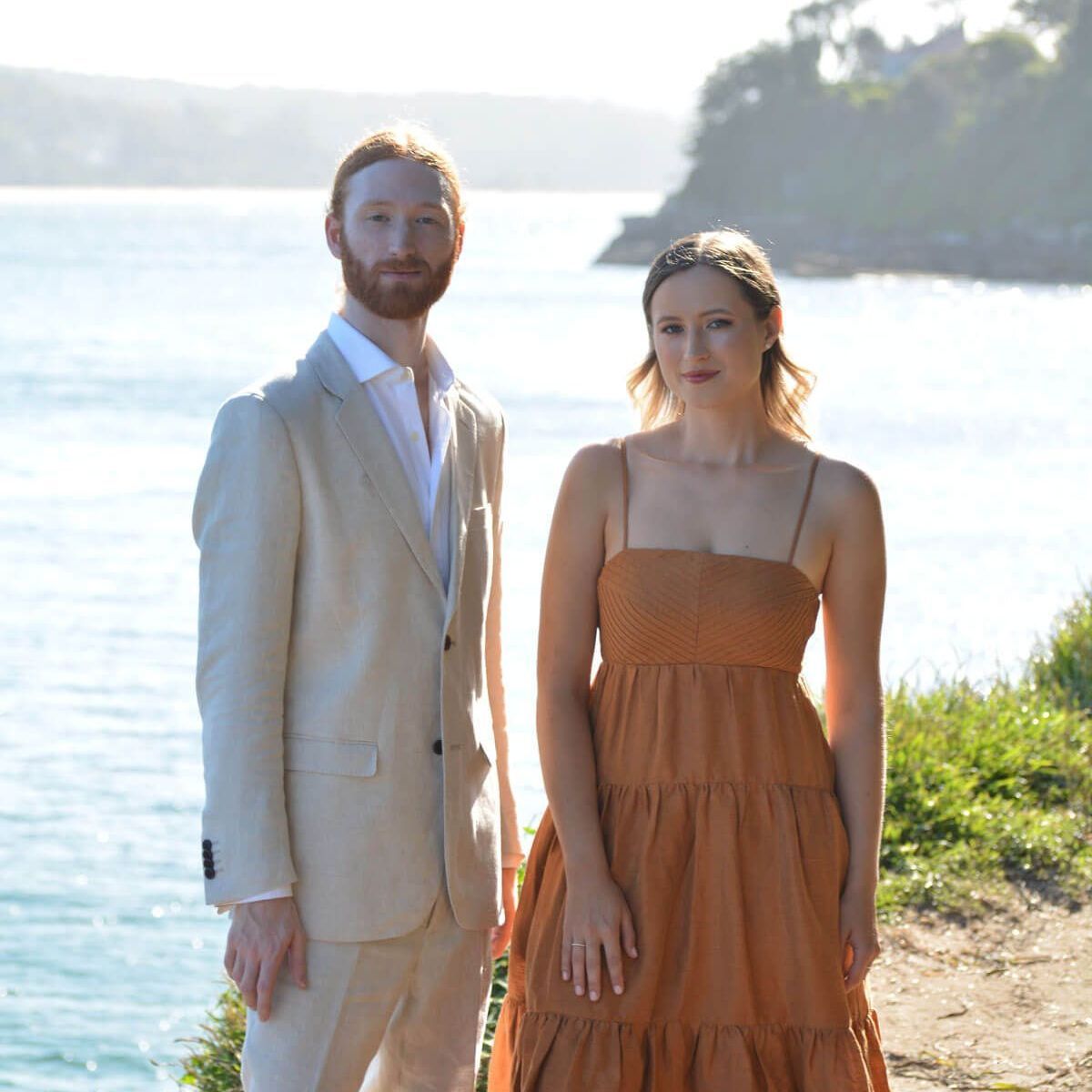 A man in a beige suit and a woman in an orange tiered dress stand side-by-side against a backdrop of water and greenery.