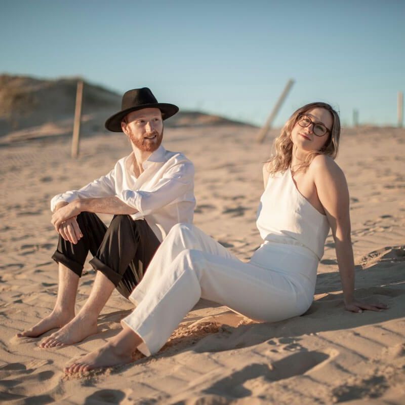 A person in a hat and white shirt and another in a white outfit sit together on a sandy beach under a clear sky.