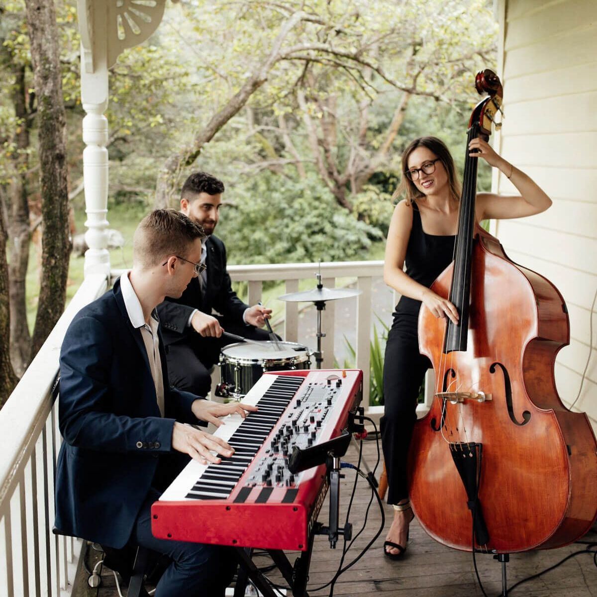 Three musicians performing on a porch: one playing a red keyboard, another drums, and a third playing an upright bass.