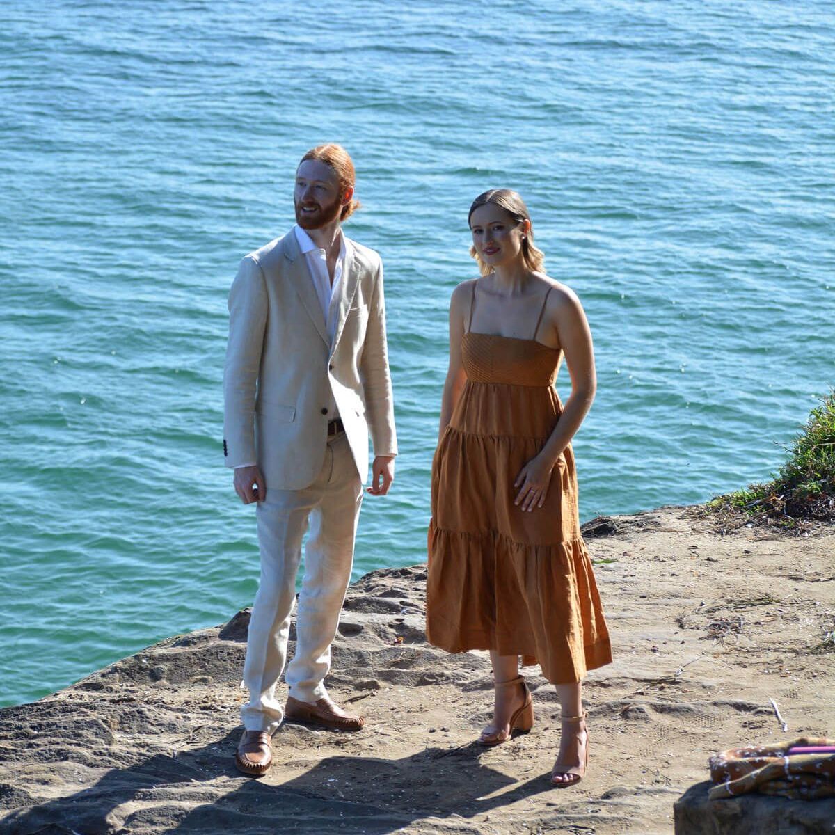 A person in a light suit and a person in an orange midi dress stand on a cliff overlooking the ocean.
