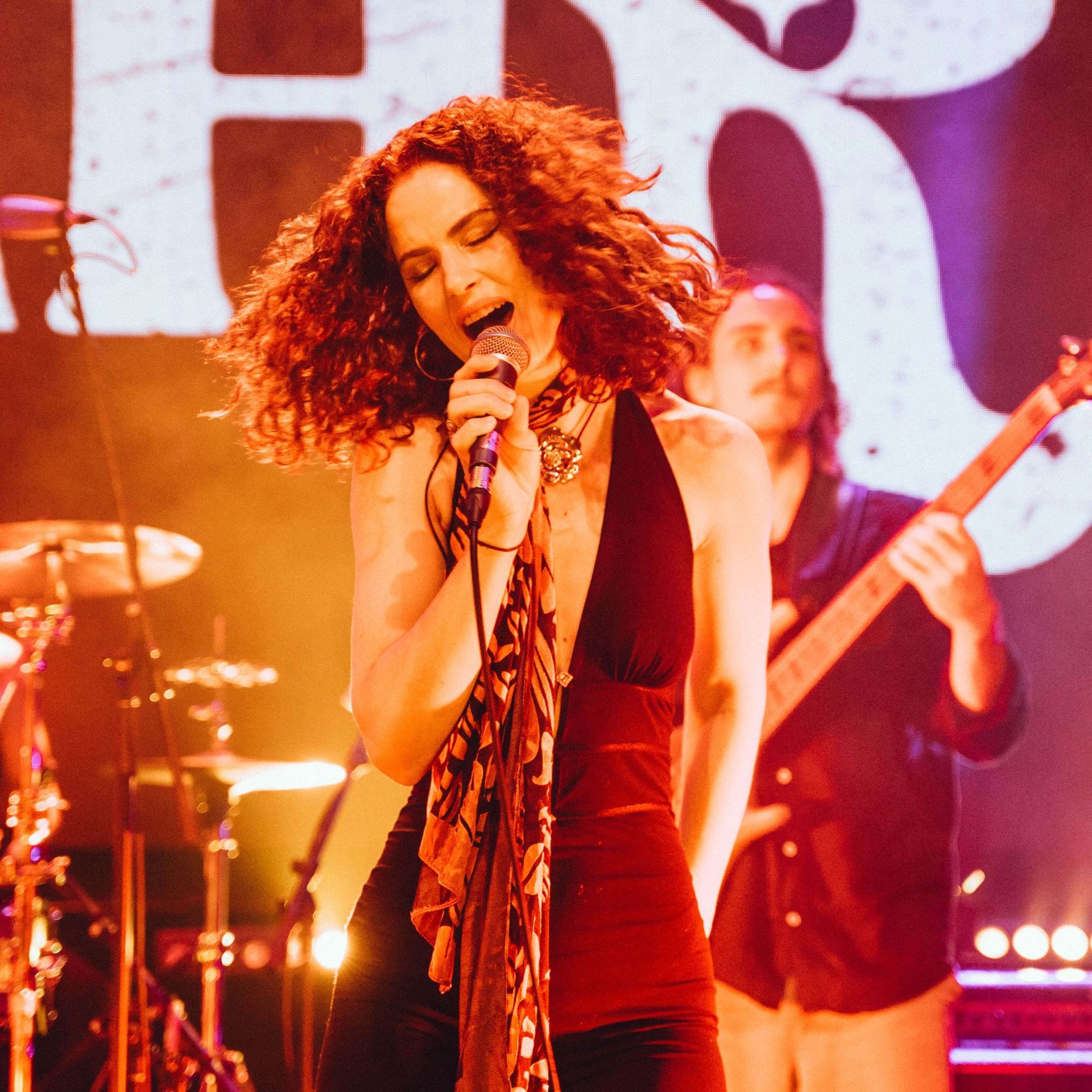A singer with curly hair performs into a microphone on stage, with a musician playing bass guitar in the background.