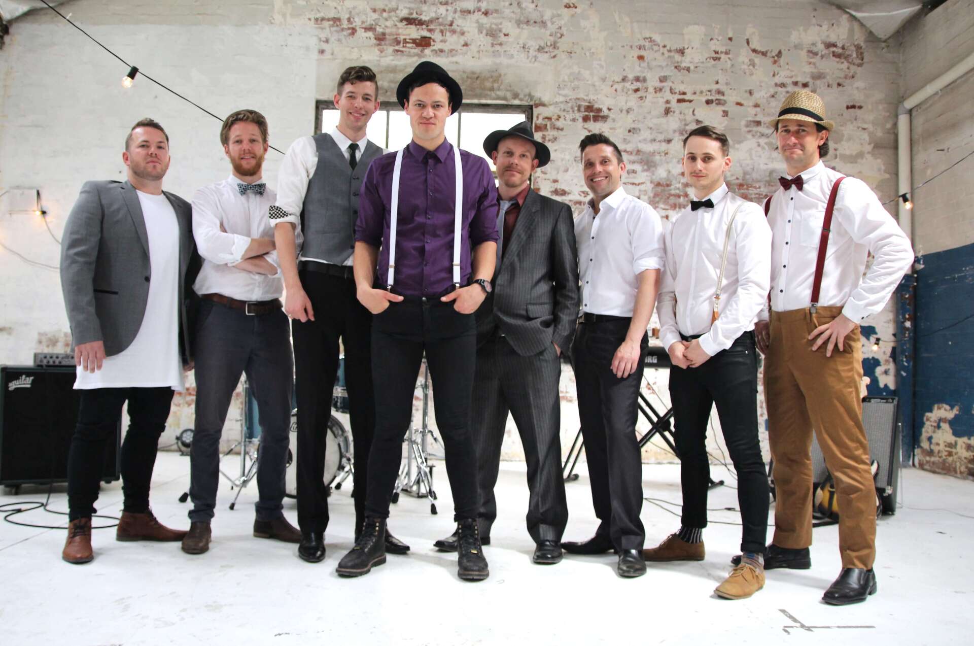 Eight men in suits and hats stand in a studio. Some have suspenders, bowties. Neutral expressions.