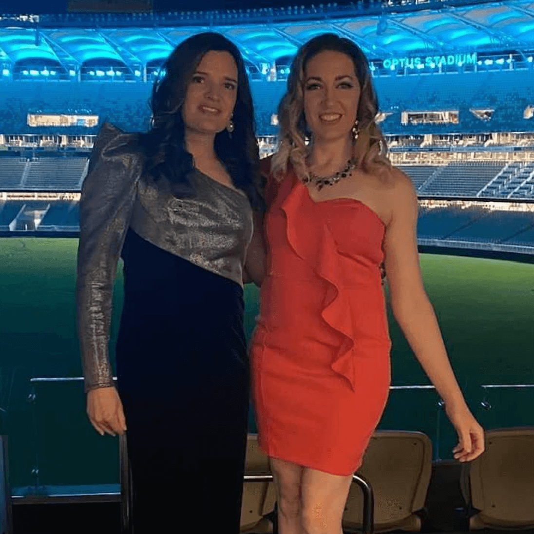 Two people pose smiling in front of the Optus Stadium field at night; one wears a metallic silver and black dress.