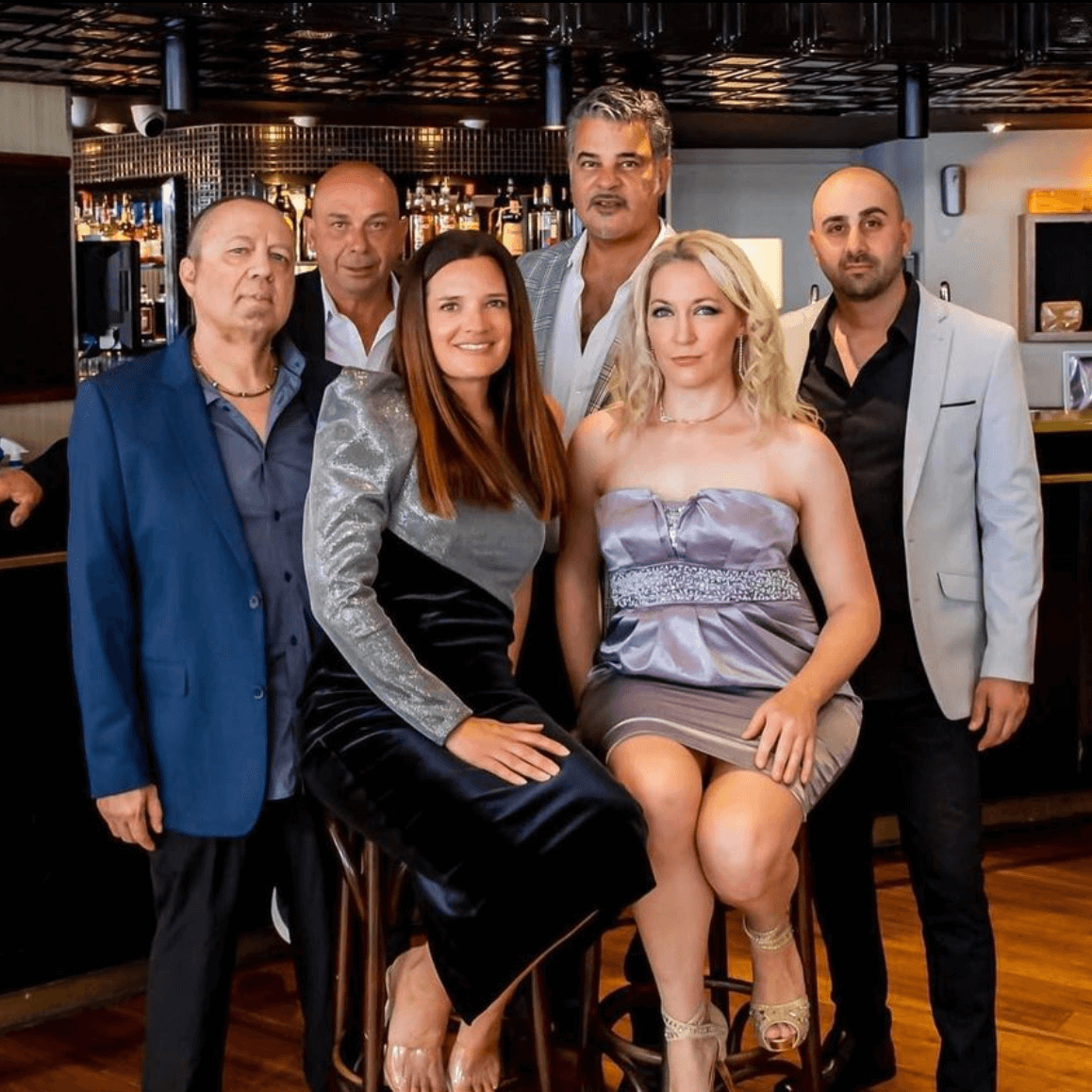 A group of six people stand and sit together for a formal portrait inside a restaurant or lounge.