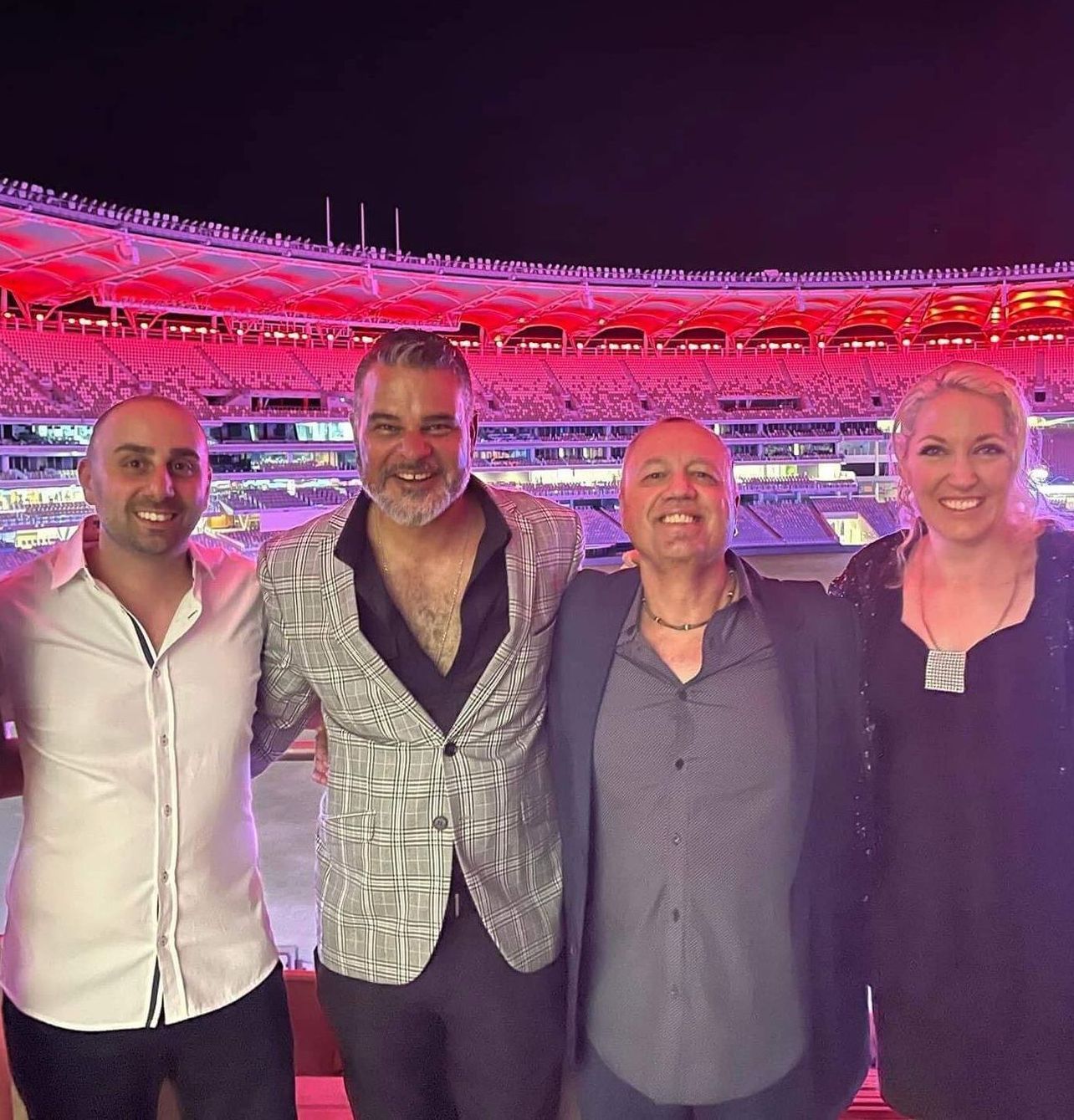 Four smiling people pose for a photo in front of a stadium lit with red lights at night.