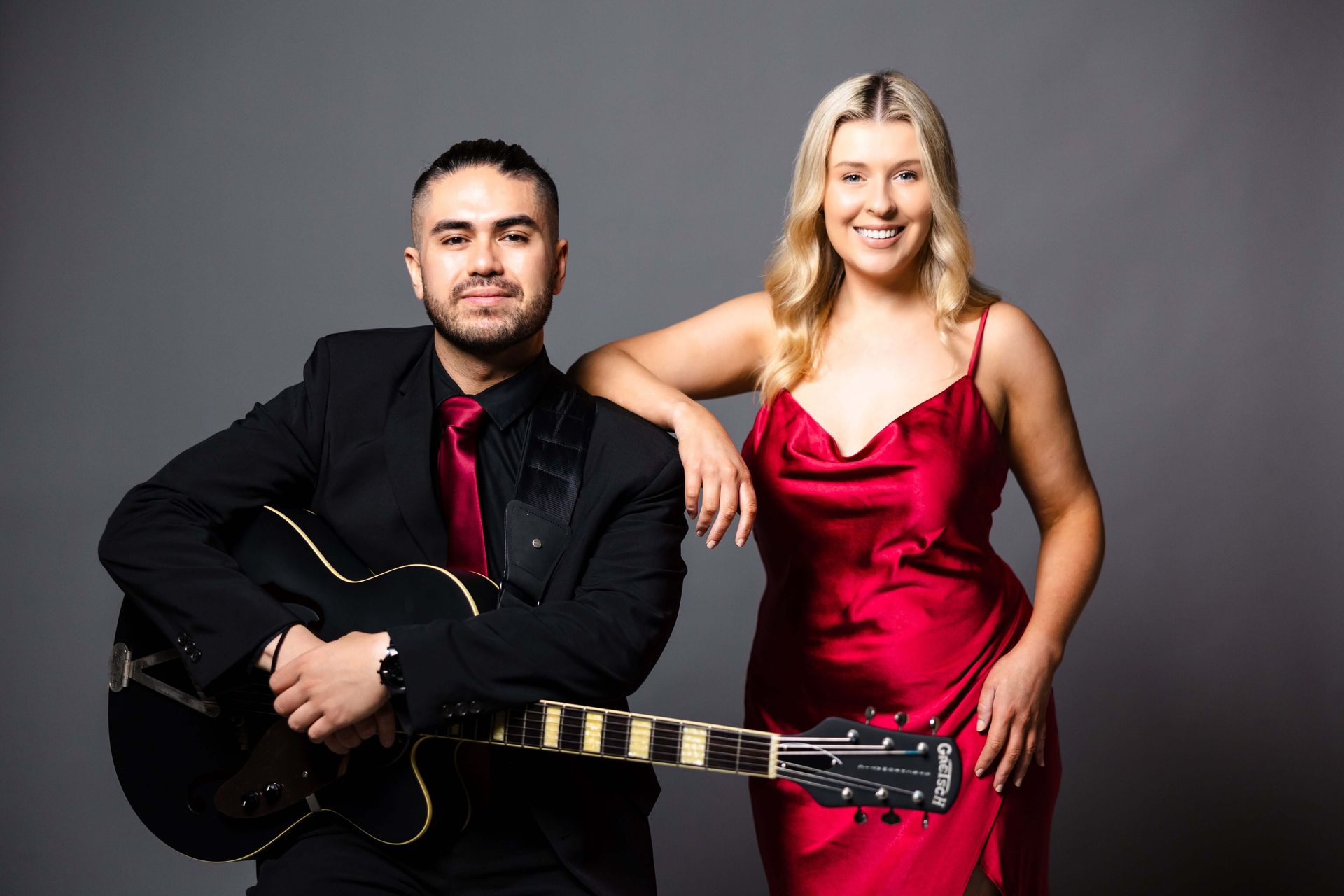 A musician in a black suit holding an electric guitar stands next to a woman in a red silk dress against a gray background.