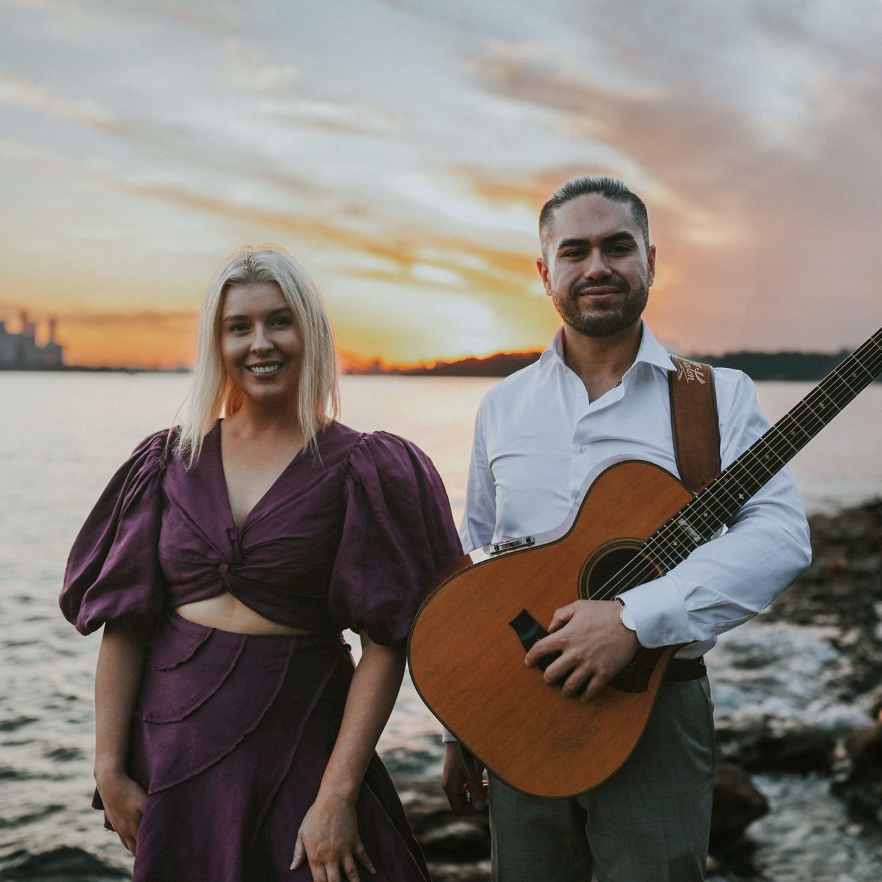 A smiling woman in a purple dress and a man holding an acoustic guitar pose together outdoors by the water at sunset.