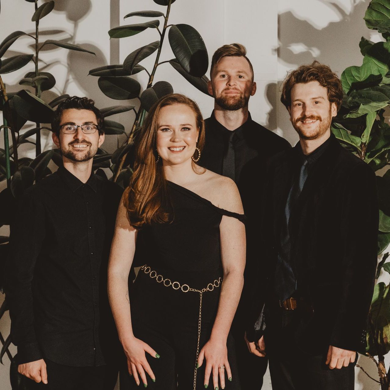 Four people in formal black attire pose in front of lush green plants.