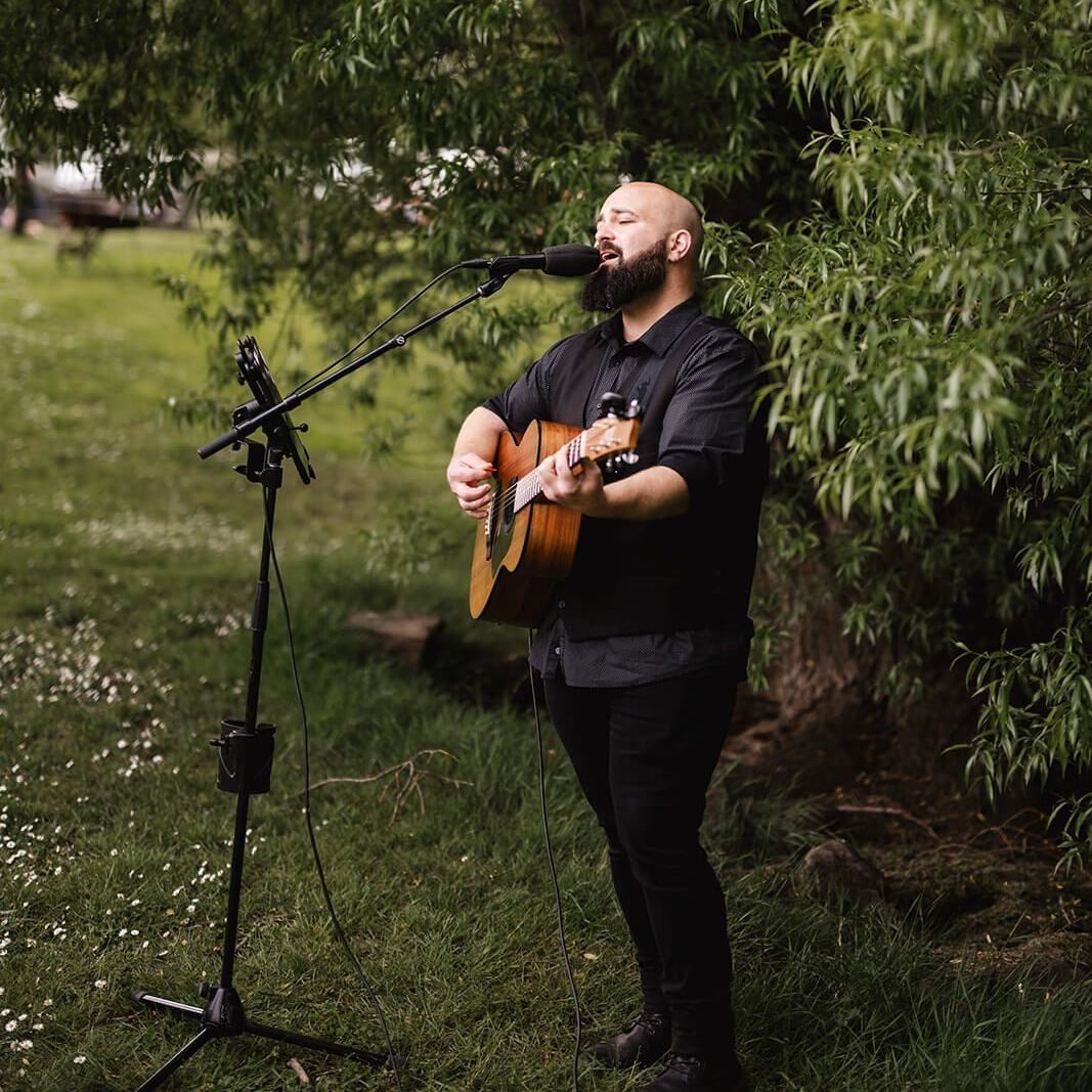 Man playing acoustic guitar and singing into a microphone outdoors on grass near green bushes