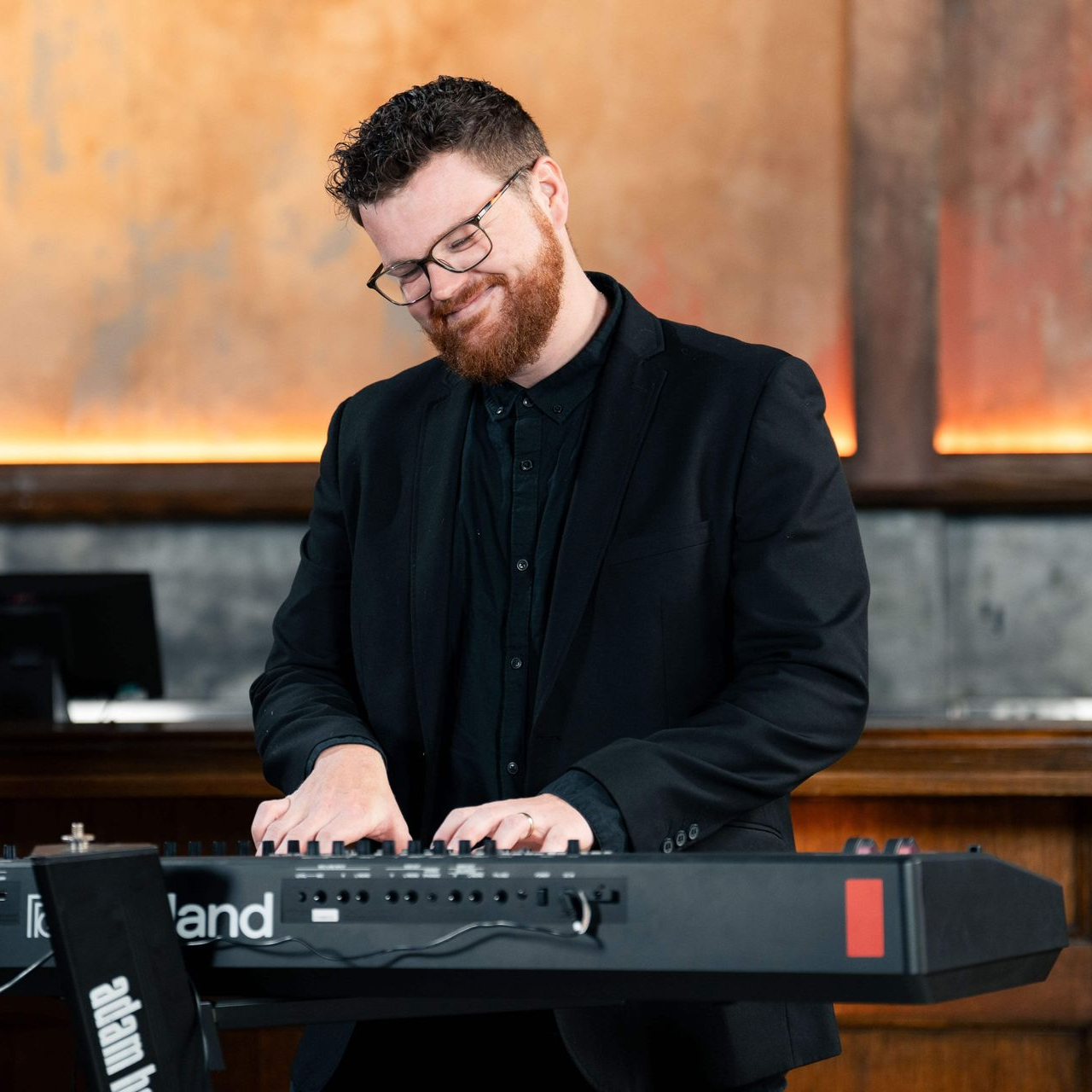 Man playing a Roland keyboard, smiling, wearing glasses and black suit.