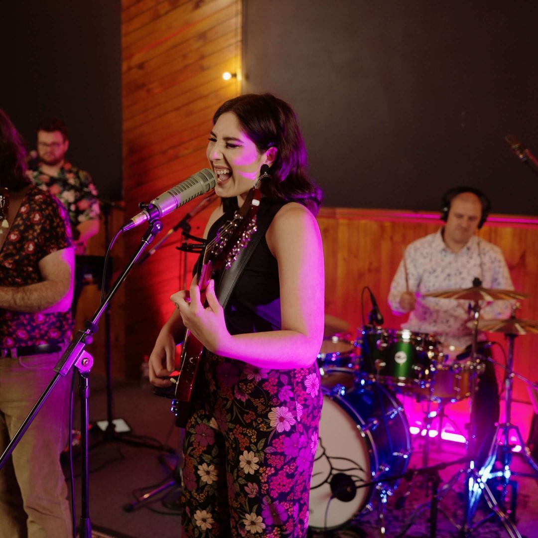 A band performing live in a studio. The singer plays guitar and sings, smiling, with a drummer in the background.