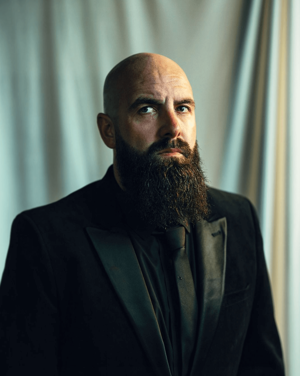 Bald man with a long beard in a black suit looking intently at the viewer.