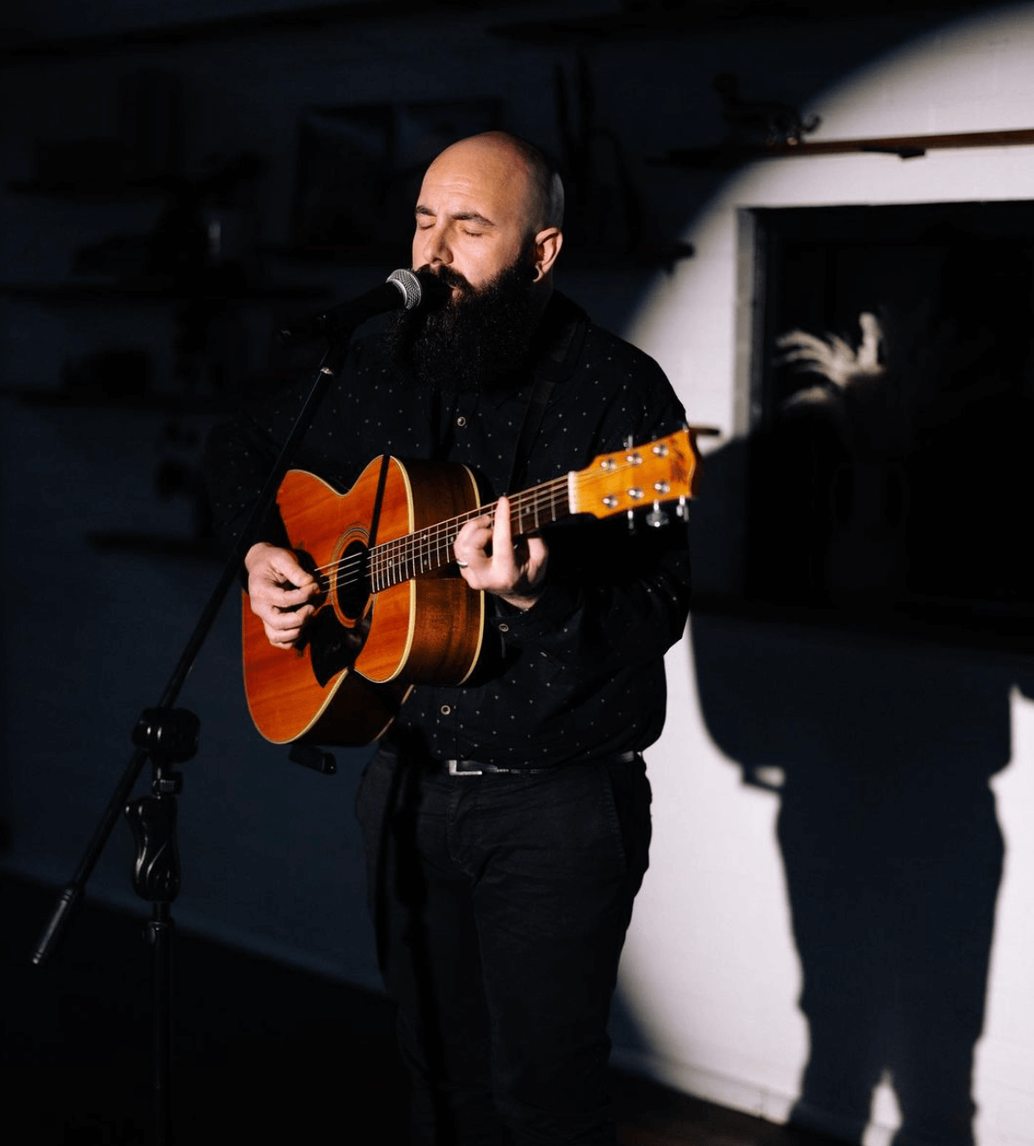 Man with beard plays acoustic guitar, sings into a microphone, stage performance.