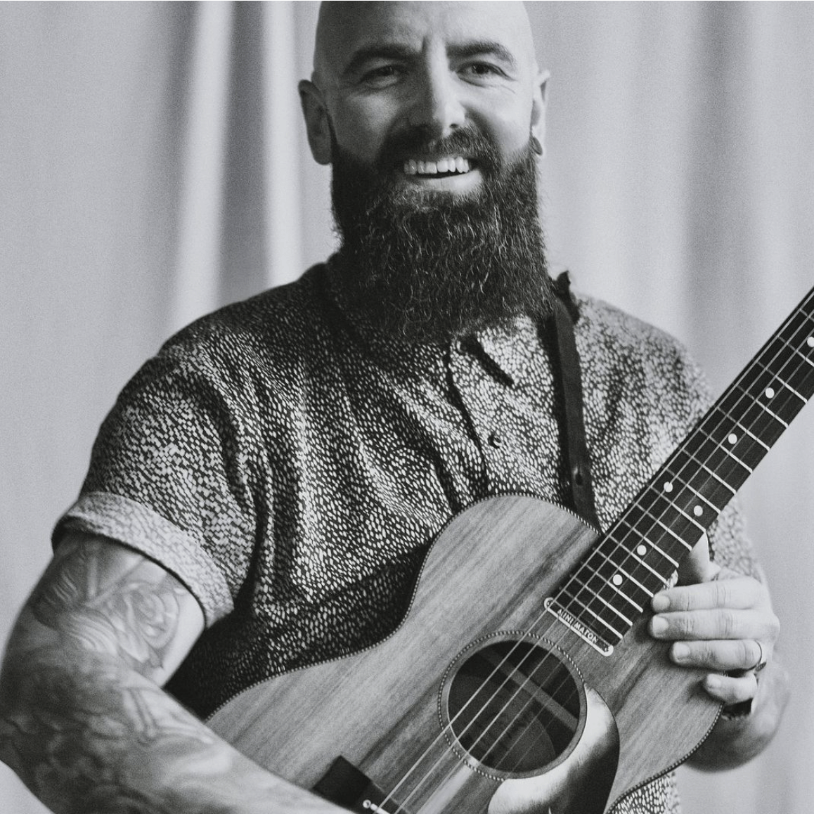 Bald man with a large beard smiles while holding a ukulele, wearing a patterned shirt; tattoo on his arm.