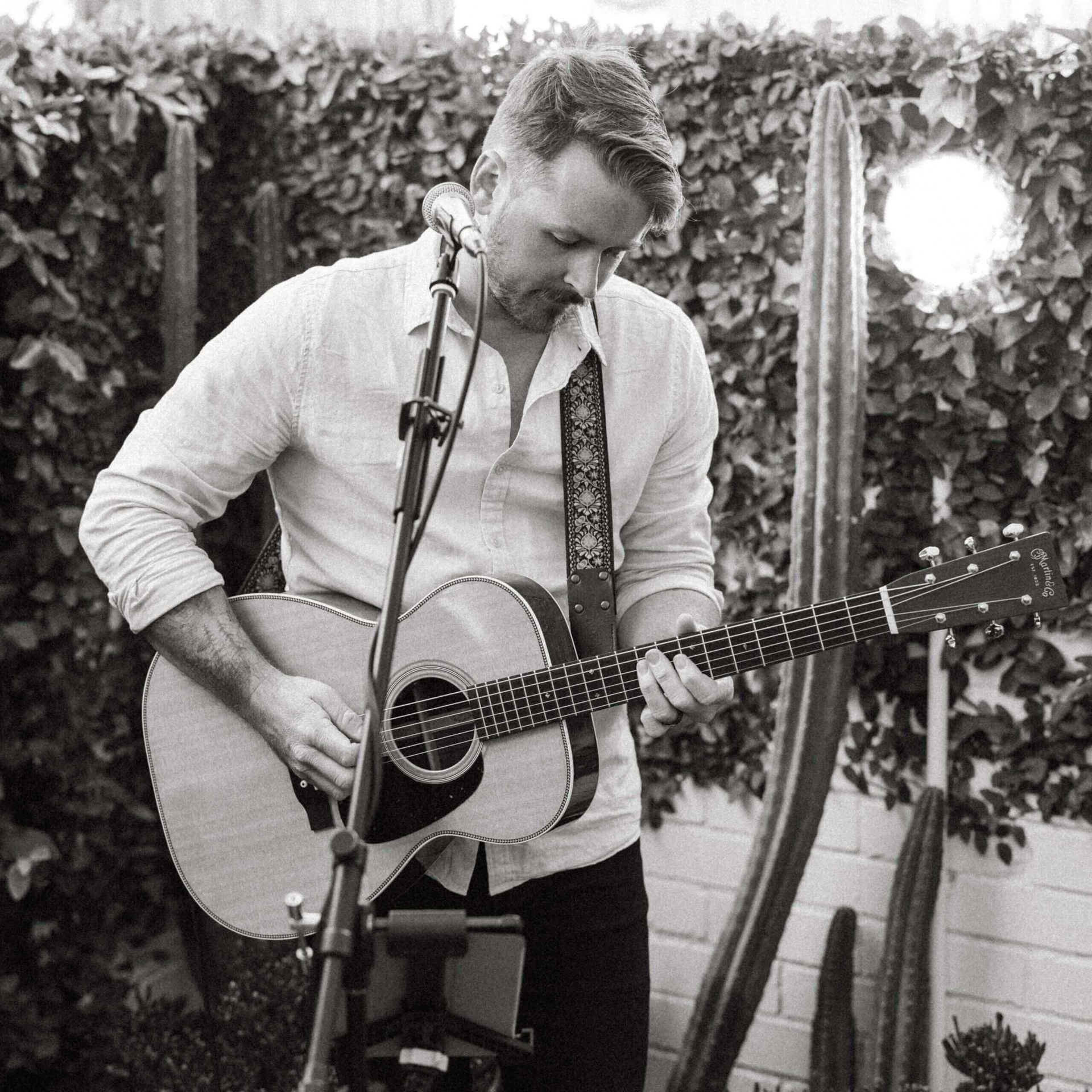 Man playing acoustic guitar, singing into a microphone outdoors; cacti and foliage in background.