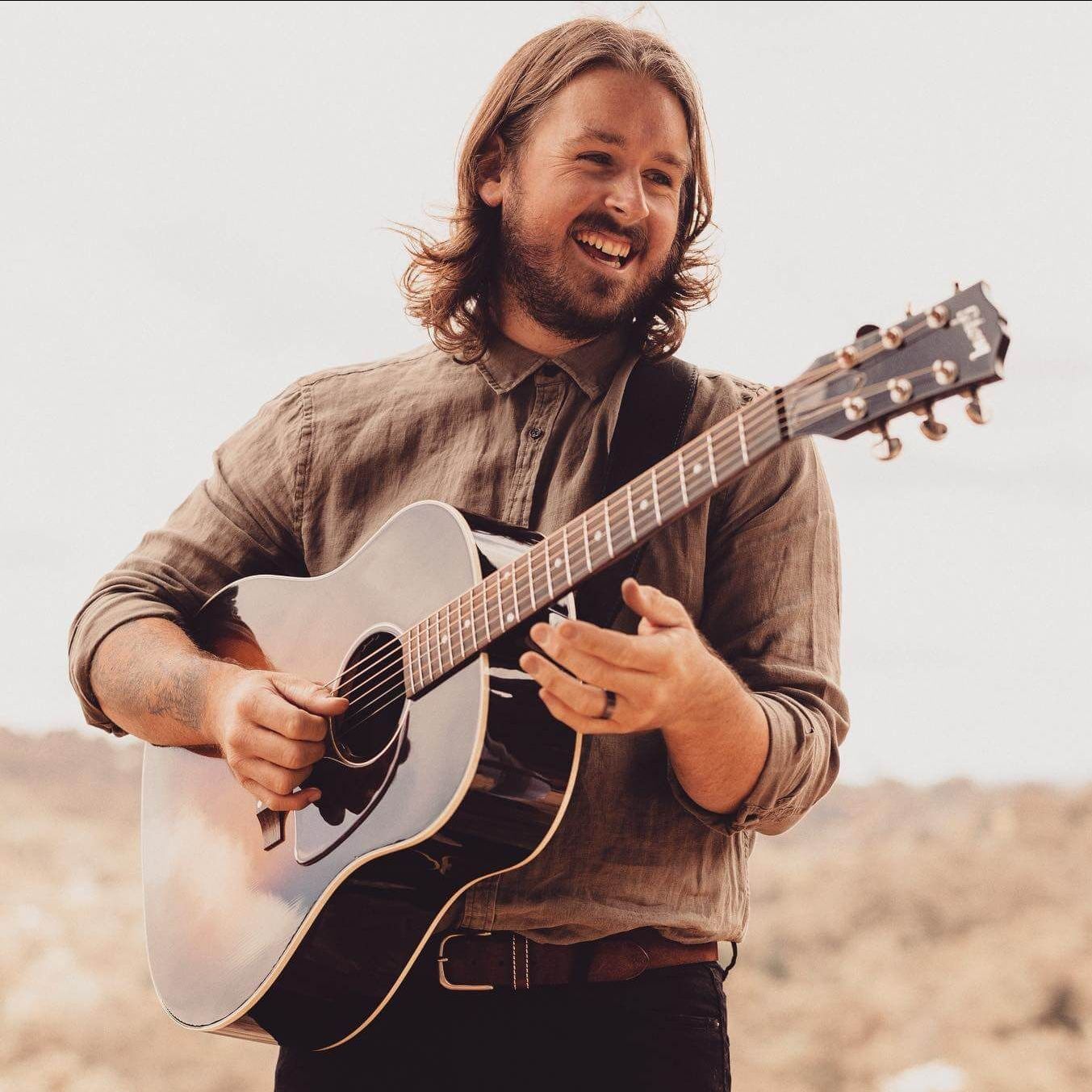 Man with long hair playing acoustic guitar outdoors, smiling. Brown shirt, guitar strap.