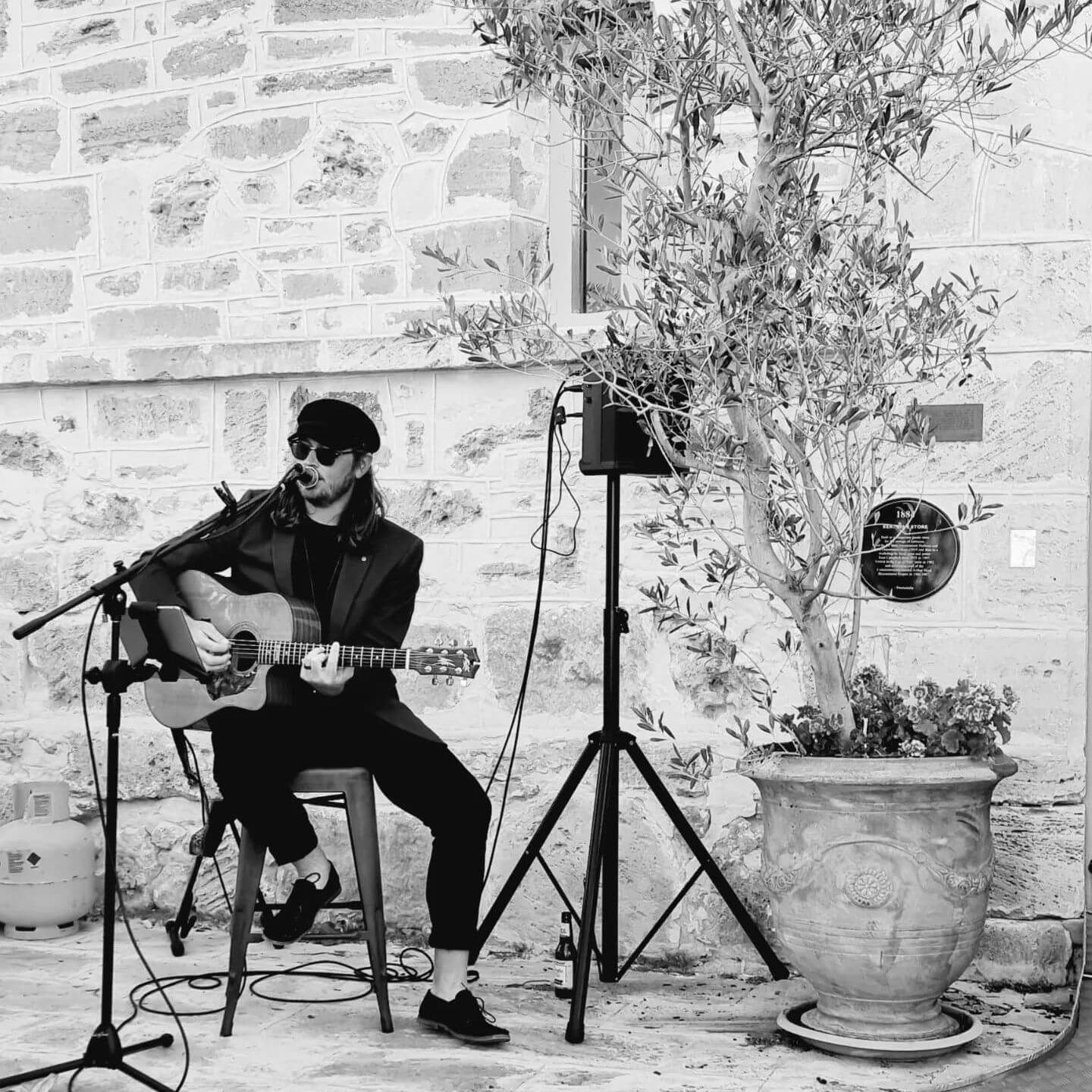 Musician playing acoustic guitar outdoors, sitting on a stool near a potted tree and stone building.