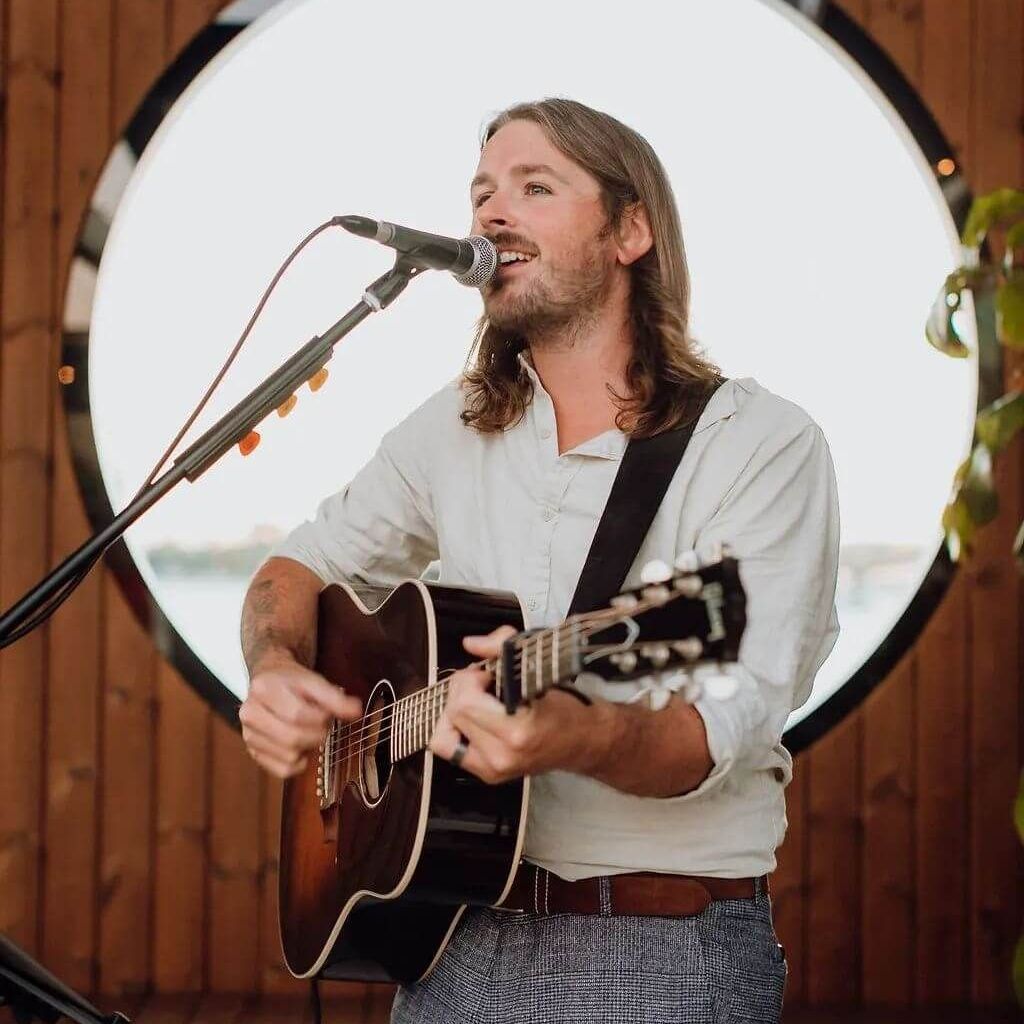 Man with long hair sings and plays acoustic guitar into a microphone in a wooden room.